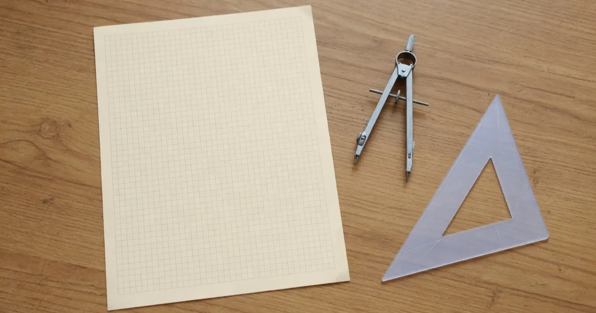 A blank sheet of pale graph paper with an empty grid, a metal drawing compass and a plain set square arranged on a drafting desk, with nothing written anywhere.